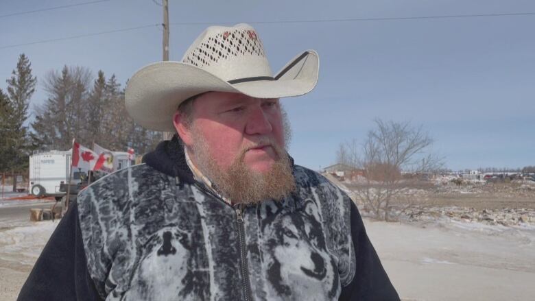 A man in a cowboy hat stands in the middle of a dirt field and looks to the side of the camera.