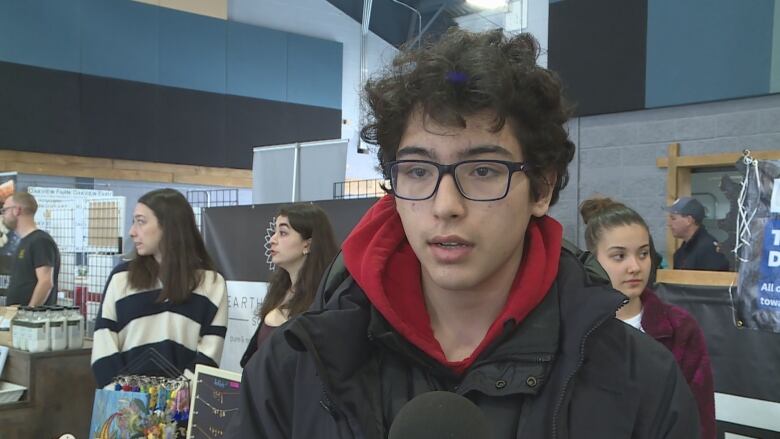 A young man in a red hoodie with a black jacket over it speaks into a microphone.