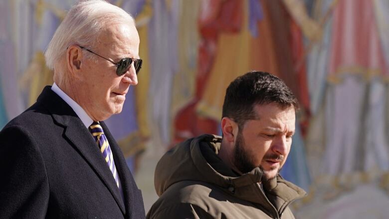 U.S President Joe Biden sports a suit and sunglasses as he walks outside alongside Ukrainian President Volodymyr Zelenskyy, wearing a brown, militaristic jacket.