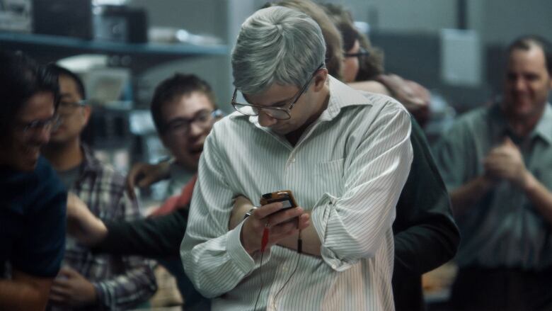 A man with grey hair and a striped dress shirt slouches and hugs his body while holding a Blackberry phone, in a crowded room.