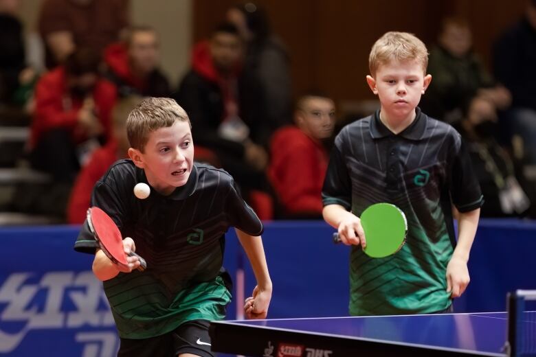 A slim boy grimaces while reaching to hit a ping pong ball, which can be seen in mid flight about 20 cm away from his red paddle. Another boy who is slightly shorter and younger looking stands to the right of him in the frame in a ready position with a green paddle held in front of his midsection.