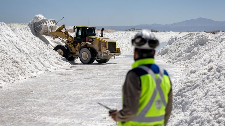 Labourer wearing flourescent safety vest stands in the foreground, looking at a dozer dumping lithium onto a piled row of the metal.