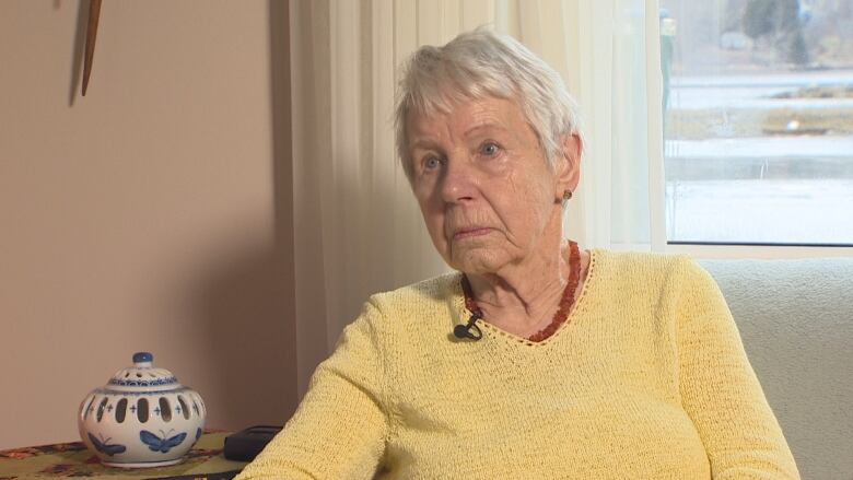 A woman in a yellow sweater sits in front of a window looking out onto a cove.