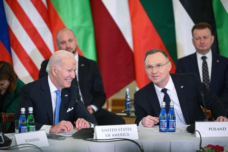 Men in suits are shown seated around a round table, with placards displaying the names of the countries they represent.