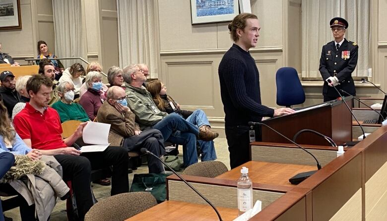 A man in a black sweater and longer hair in a bun stands at a microphone and podium, speaking to councillors seated to the right and a public gallery full of people visible to the left.