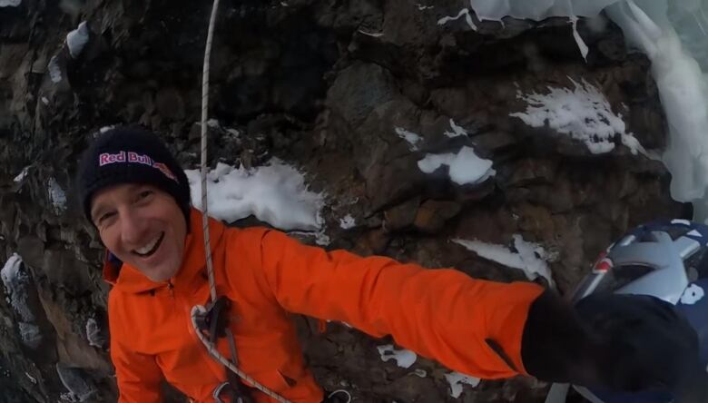 A man with toque and in jacket lies on a rock slope with a cable on his body.