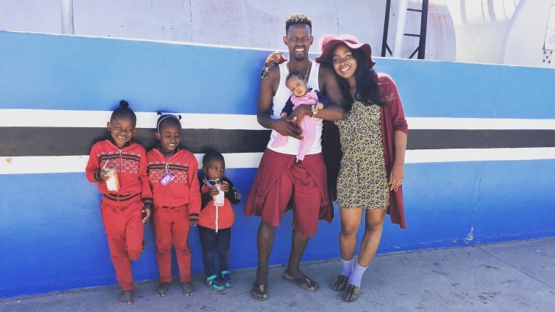 A woman wearing a short dress and sunhat poses in front of a Botswana flag mural with three children and a man holding a baby. 