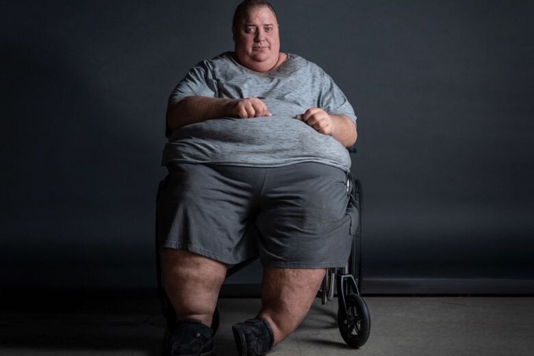 Wearing a prosthetic body suit and dressed as his character in The Whale, Brendan Fraser sits in a wheelchair against a grey background.