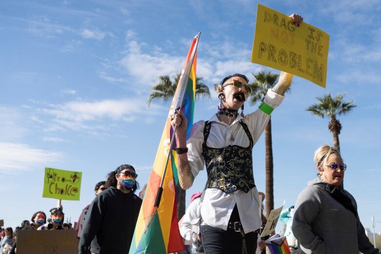 A person at a protest holds a pride flag in one hand, and holds up a sign with the other that reads: 