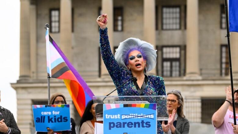 A drag queen raises her fist in the air at a podium.