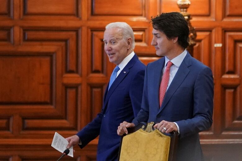 Two men in suits walk together. The older one, with white hair and a blue tie (Joe Biden) looks mid-sentence. The younger one, with touselled brown hair and a red tie (Justin Trudeau) raises his eyebrows.