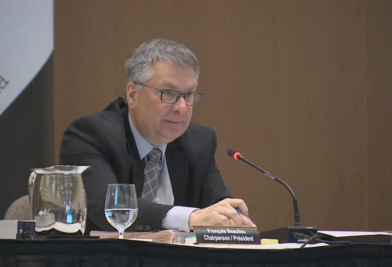 A man in a suit sits at a table with a microphone infront of him. 