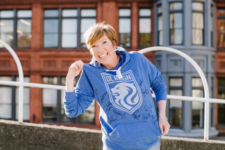 A woman with short, reddish hair shows off an autographed Ol Reign soccer team hoodie she is wearing. 