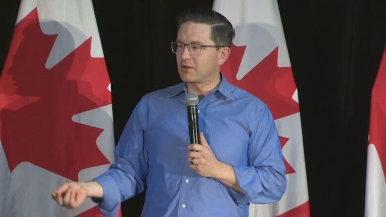 A man wearing a blue shirt holding a microphone and standing on a stage. Behind him is a black backdrop and Canadian flags. 