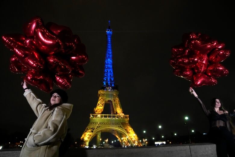 Two woemn hold red bunches of balloons in front of the illuminated structure.