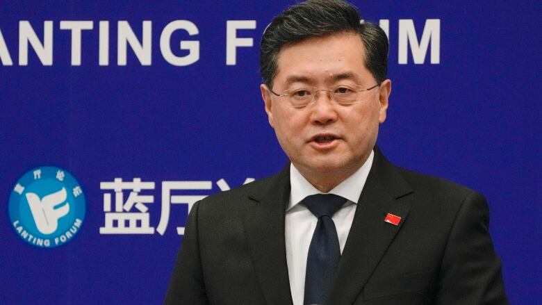 A dark haired man with galsses stands at a podium in front of a blue wall with Chinese characters on it