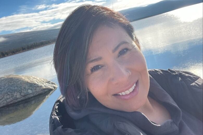 A selfie of a dark-haired woman smiling outdoors by a lake.