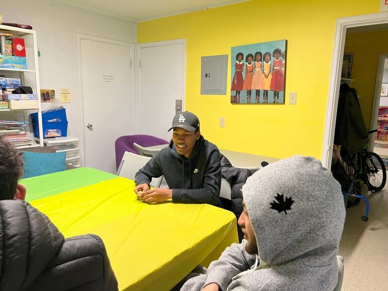 Armon Jeffries ats at a yellow table, wearing all black, including a black hat. His friend Tayon Briand (left) is wearing a black winter coat and Mikell Logan (right) is wearing a grey hoodie with his arms crossed.