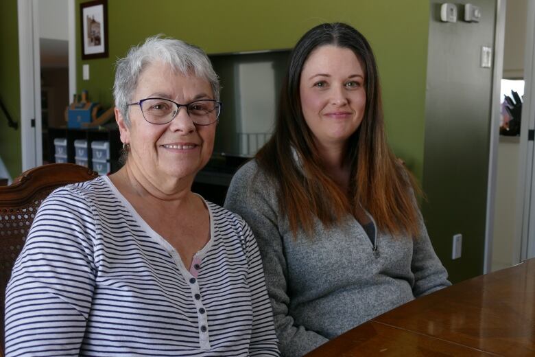 An woman with white hair and glasses wearing a black and white striped shirt sits next to a woman with long brown hair wearing a grey sweater smile as they sit at a dining room table.