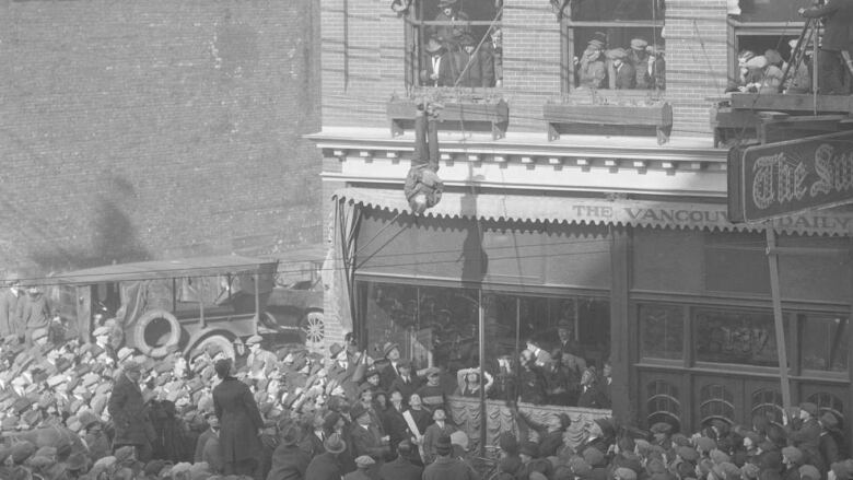 A crowd of people forms around the Vancouver Sun Building, Houdini is suspended upsidedown middair in a straightjacket. 