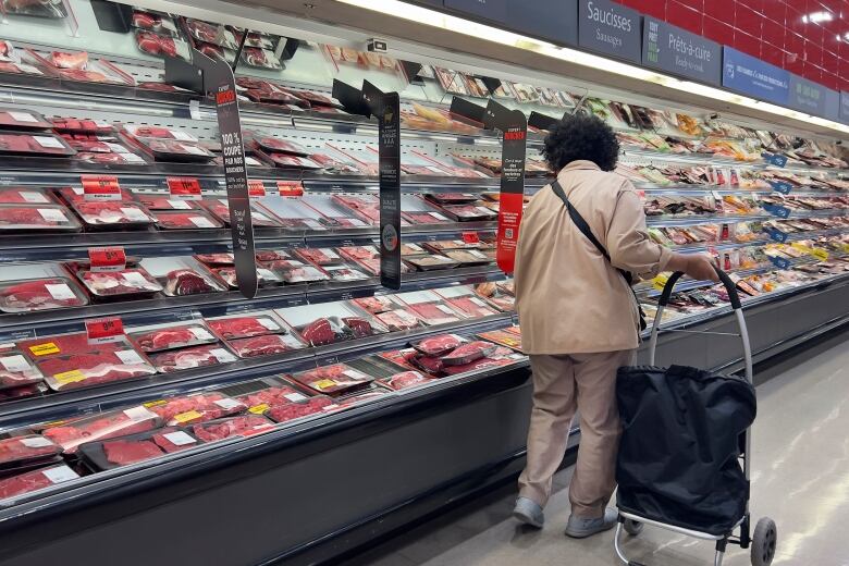 A customer in a brown jacket looks at meat in a grocery store aisle.