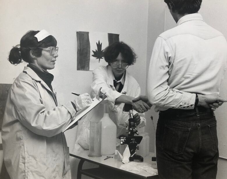 One woman in a lab coat takes notes, while another rests her arms on a microscope, as a man, seen only from behind, stands still in the foreground,.