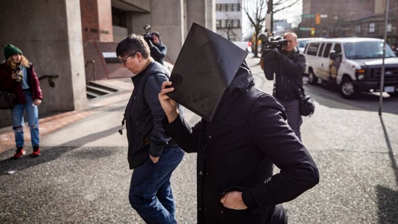 A woman, surrounded by news camera, obscures her face with a file folder as she enters a courthouse.