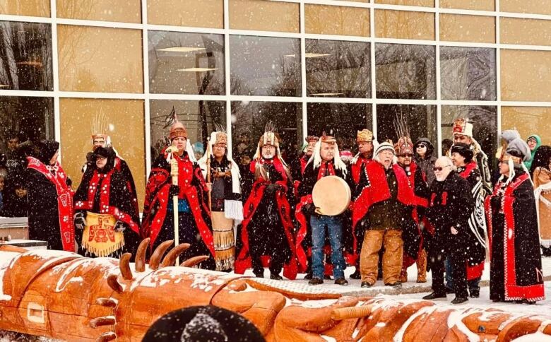 A group of people in Indigenous clothing are holding drums in front of a building and behind a laid-down totem pole.