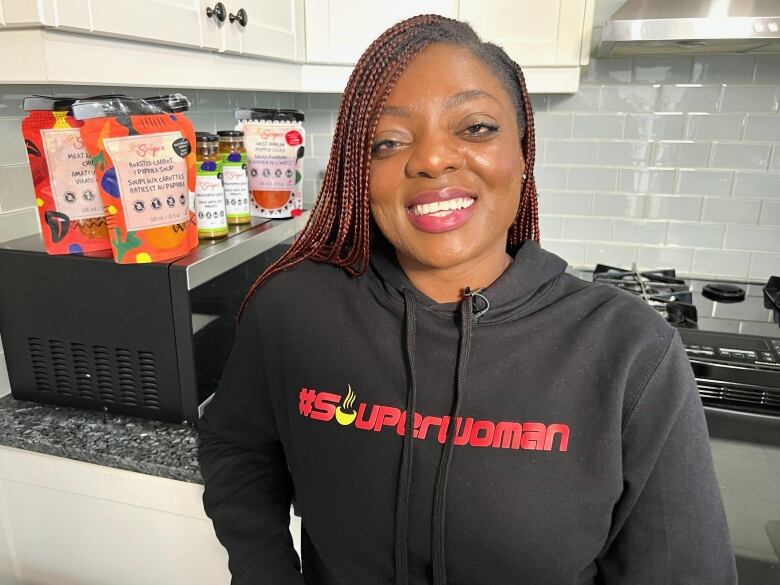 A woman, Lola Adeyemi, is shown smiling in front of products that her food company, It's Souper, sells.