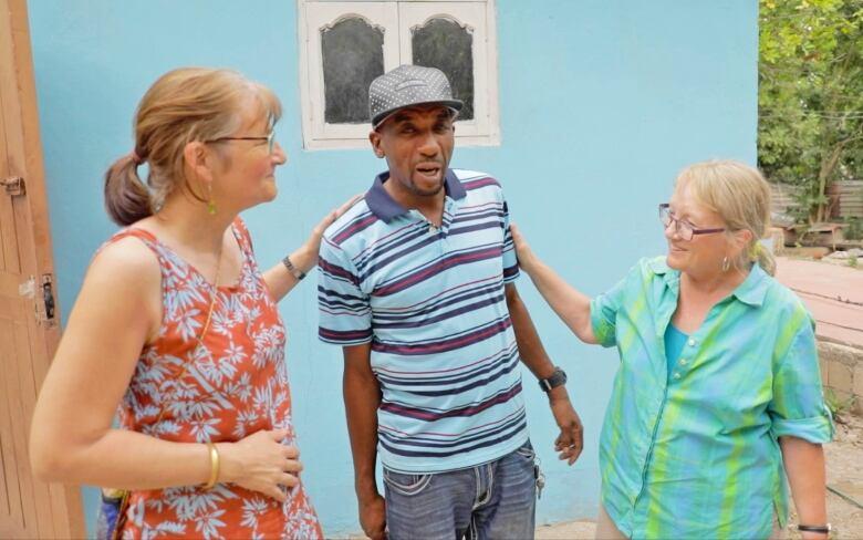 Two women and a man standing and chatting, the two women have one hand on the man's shoulders.