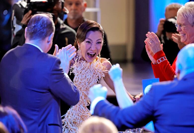 A woman in a gold and black dress smiles as people applaud her.