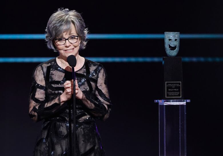 A woman with grey hair and wearing a black dress stands at a microphone with her hands clasped in front of her.