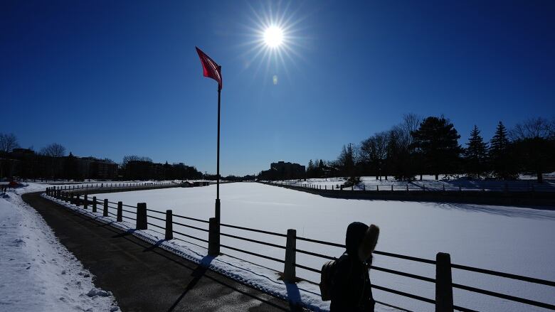 A person, silhouetted by the sun, walks on a cement path adjacent to a snow-covered canal.