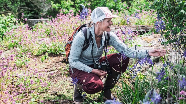 A blonde woman wearing outdoor gear, crouches down to look at plants in a lush forest.