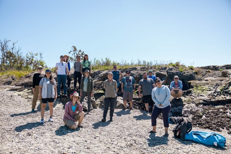 A group of people wearing outdoor clothing pose for a photo on a pebbly and rocky beach.