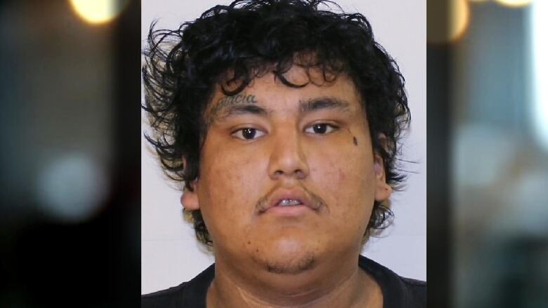 A man with dark hair, dark eyes and a tattoo over his right eyebrow poses in a police photo.