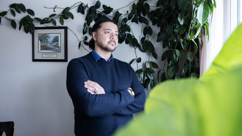 A man with black hair wearing a blue sweater stands with his arms crossed, looking to the side out a window. Leaves from a plant frame the photo.