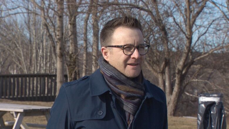 A white man with glasses and short hair stands outside in a navy buttoned coat and scarf. He looks off to the right of the frame, while bare trees and a picnic table can be seen behind him