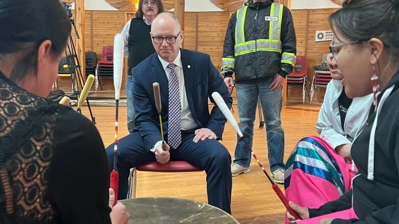 A bald man in glasses, a navy suit and striped tie takes part in a drumming circle. He holds a drum stick while he is flanked by others doing the same.