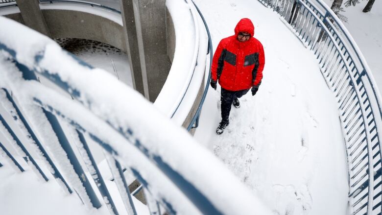 A man in a red jacket walks along a snow-covered concrete walkway with metal side railings. 