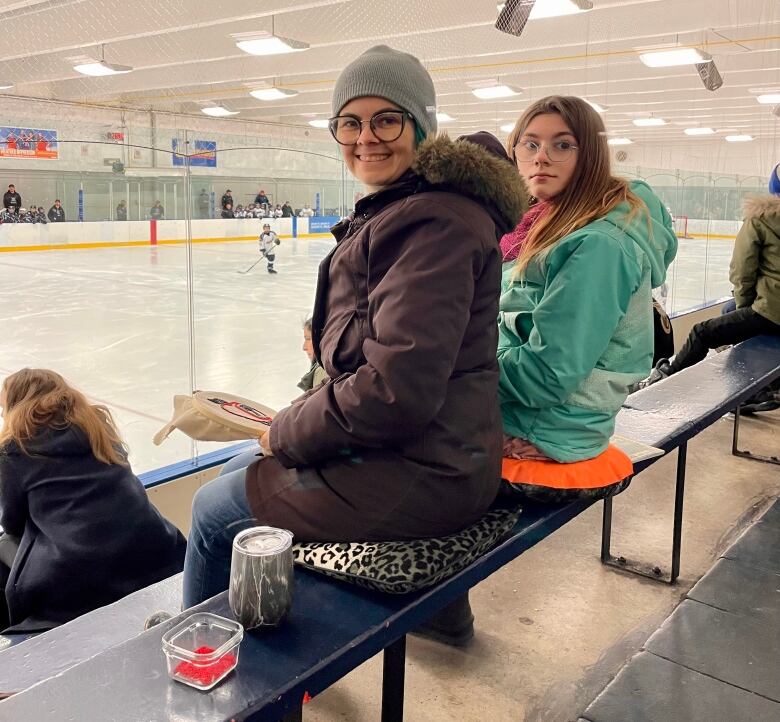 A woman and girl, both wearing winter coats, sit on cushions on the wooden bench inside a hockey arena. 