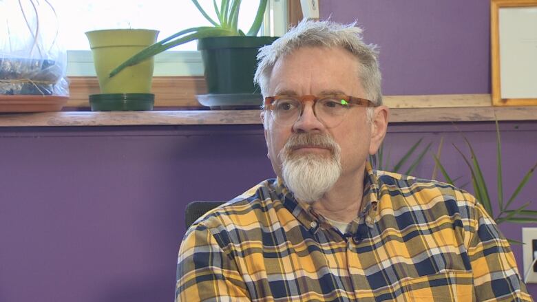 A man with short grey hair and tortoise shell glasses wears a blue, white and yellow plaid shirt. He is sitting in front of a purple wall with shelves.