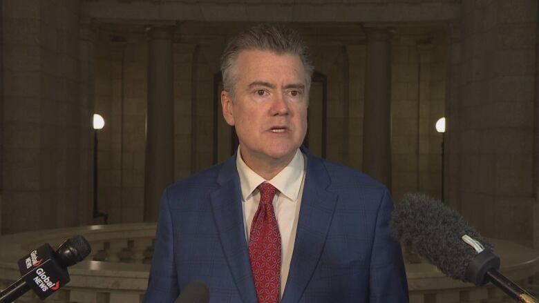 A man with grey hair wearing a blue suit and a red tie stands behind microphones.