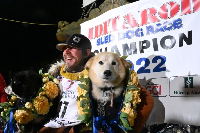 A man sits with his arm around a dog in front of a sign reading, 'Iditarod sled dog race champion 2022.'