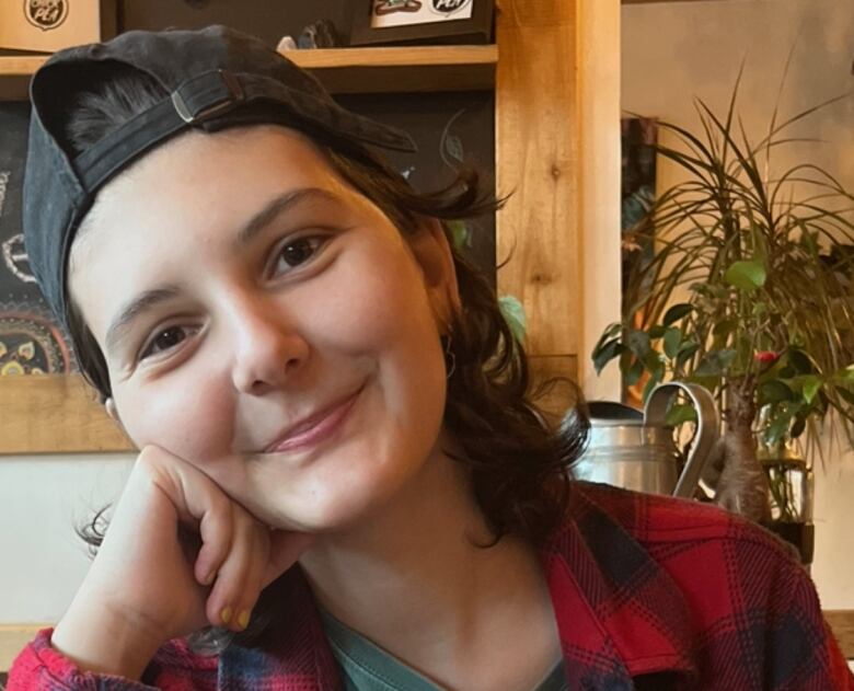 A woman wearing a baseball cap backwards and a red flannel shirt poses in front of a wooden shelf.