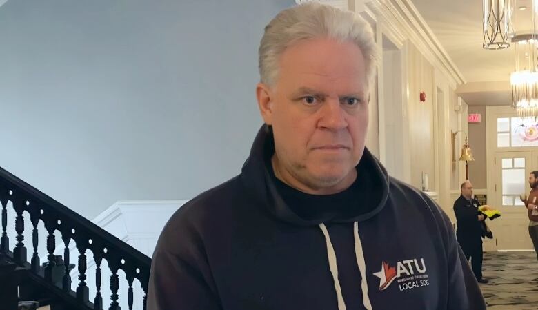 A white man with white hair looks off to the side of the frame with a stern expression, standing in a City Hall hallway.