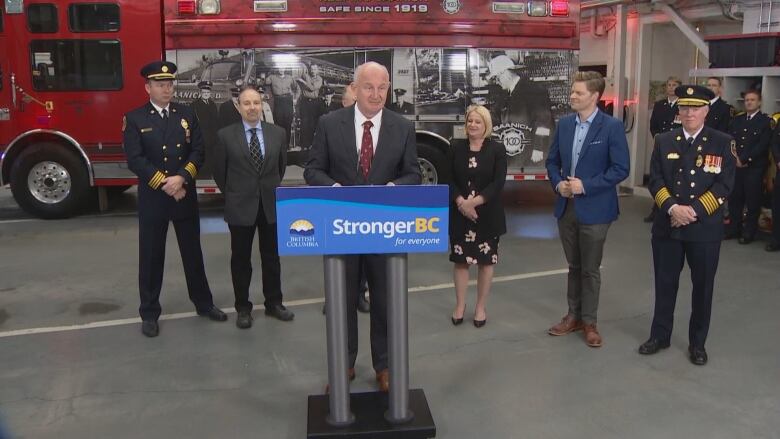 Minister Mike Farnworth is at a podium speaking. In the background is a firetruck and other guests and ministers for the announcement.