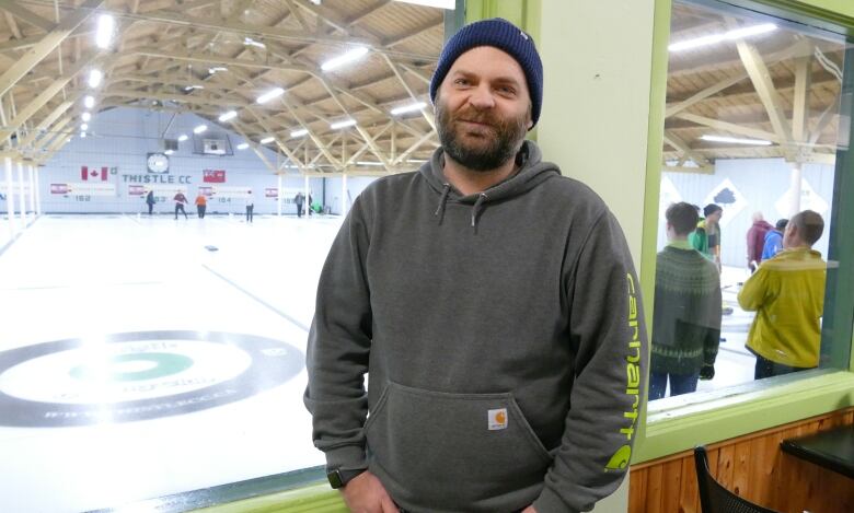 A bearded man in a tuque stands in front of four sheets of curling ice.