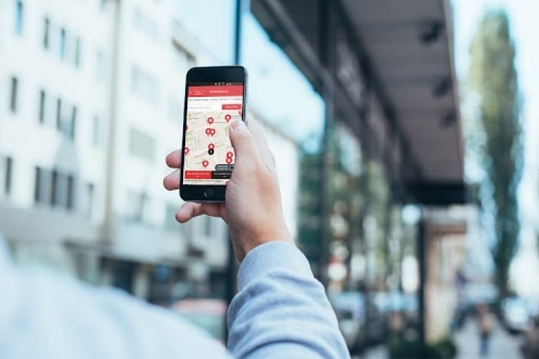 A person in a grey sweater holds up a cell phone with the Go Here bathroom app open, showing the nearest available public washroom.