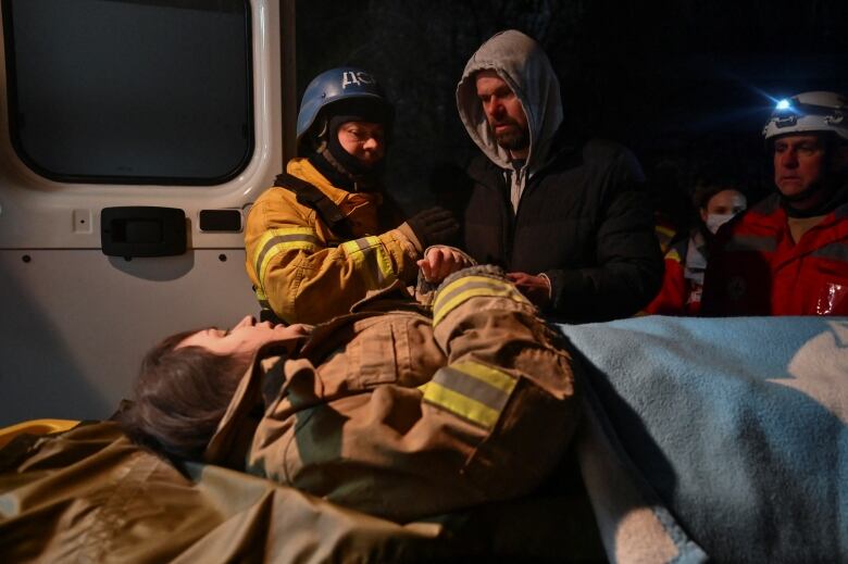 A bearded man dressed in a grey hoodie and jacket looks down at a woman who is lying on a stretcher.